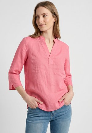 Mujer con cabello castaño claro lleva una blusa rosa de manga larga y vaqueros azules, con las manos en los bolsillos, mirando ligeramente hacia la izquierda sobre un fondo blanco.