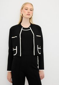 Veste noire courte avec bord blanc et deux poches avant, portée avec un haut noir. Présente des accents dorés et une texture lisse.