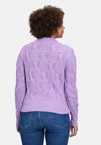 Lila, gestrickter Pullover mit einem Zopfmuster, geripptem Saum und Bündchen sowie einem hohen Kragen. Trägt eine Rückansicht mit blauen Jeans.