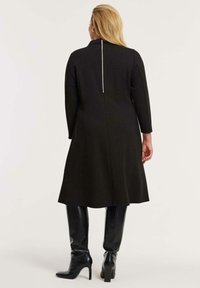 Schwarzes knielanges Kleid mit einem Reißverschluss auf der Rückseite, langen Ärmeln und einer ausgestellten Silhouette; kombiniert mit glänzenden schwarzen kniehohen Stiefeln.