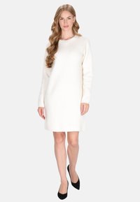 Frau mit langen, welligen Haaren, die ein weißes, langärmeliges Strickkleid und schwarze High Heels trägt, vor einem hellgrauen Hintergrund steht.