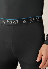 Sorte aktive leggings med en glat tekstur, der har en bred elastisk talje med mærket "NEXT ACTIVE" og blå detaljer.