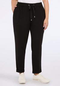 Person i svarta joggerbyxor med dragsko och vita sneakers, stående med en hand i fickan mot en enkel bakgrund.