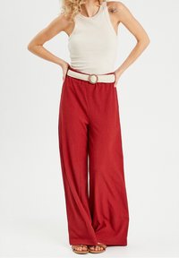 Pantalon rouge à jambes larges en tissu texturé, doté d'une ceinture blanche tressée avec une boucle circulaire. Porté avec un débardeur crème et des sandales.