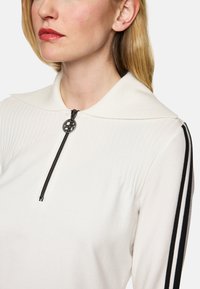 Weißer Zip-Hoodie mit großem Kragen, strukturierten vertikalen Streifen und schwarzen Seitenelementen. Verfügt über einen runden Reißverschluss mit einem Logo.