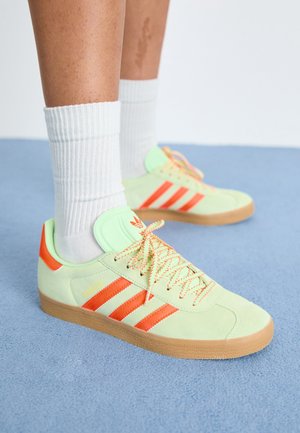 Fødder iført hvide ribstrikkede crew-strømper og blege grønne Adidas-sneakers med orange striber og gummisåler på et blåt tæppe.