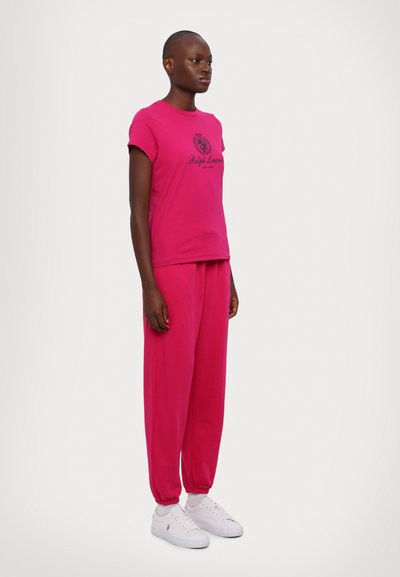 T-shirt de algodão rosa com mangas curtas e logotipo preto, combinado com calças de moletom rosa de corte solto e ténis brancos. Textura suave.