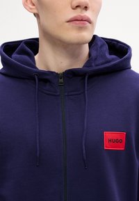 Marineblå hettegenser med teksturert innside, frontglidelås og rød brodert logopatch. Har snor i hetten og kengurulomme.