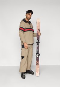 Μπεζ σκι jacket με κόκκινες και μαύρες ρίγες, μπεζ cargo παντελόνι με μαύρες λεπτομέρειες, κρατώντας ένα σκι με ανοιχτά και σκούρα σχέδια. Μαύρες μπότες σκι.