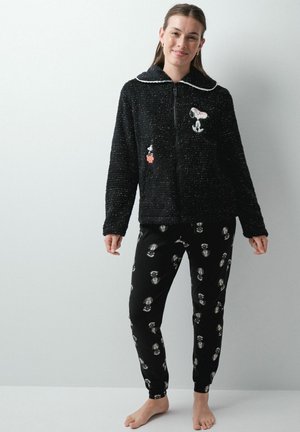 Jeune femme portant une veste zippée duveteuse noire à thème Snoopy et un pantalon assorti, debout pieds nus devant un fond clair uni.
