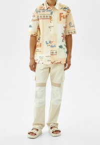 Camisa de manga corta color crema con estampados multicolores, combinada con pantalones cargo beige que presentan acentos de malla y una cinturilla ancha. Se lleva con sandalias blancas.