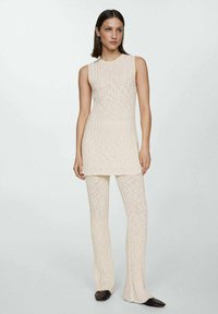 Kvinna klädd i en krämfärgad ärmlös långstickad topp och matchande texturerade stickade byxor med svarta spetsiga skor, stående mot en ljus bakgrund.