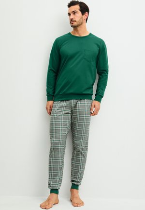 Mann mit grünem Langarmshirt und grauen karierten Pyjamahosen steht barfuß an einer weißen Wand.
