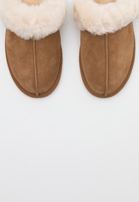 Paire de chaussons en daim marron avec doublure en laine crème duveteuse sur fond blanc.