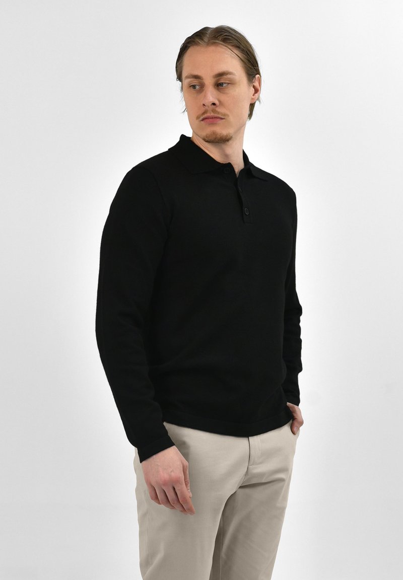 Zwarte polo met lange mouwen en kraag, met een knoopsluiting aan de voorkant en geribbelde manchetten. Draagt men met lichtbeige broek.