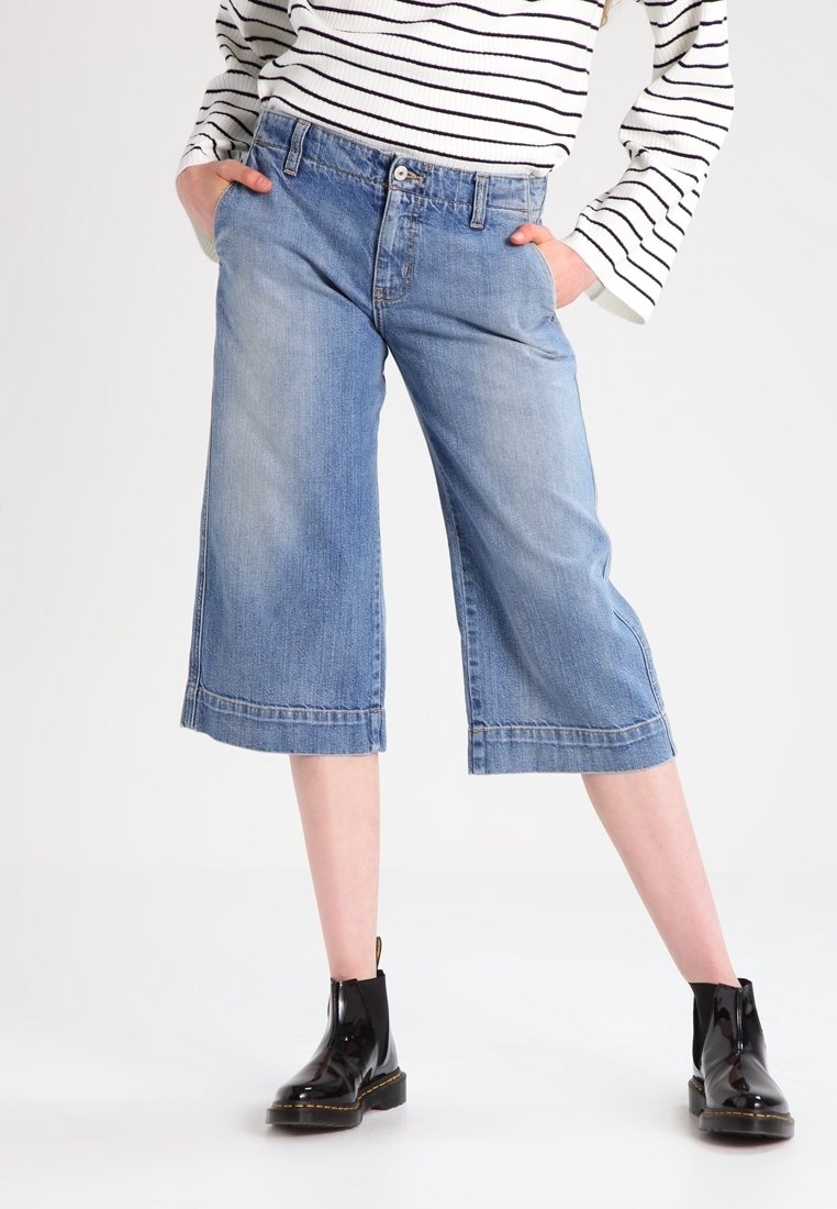 Personne portant un haut rayé à manches longues, un short en jean bleu à jambes larges arrivant aux genoux, et des bottines noires à talon bas, mains dans les poches.