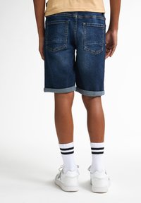 Shorts in denim scuro con risvolti, dotati di tasche posteriori e una texture liscia. Indossati con calzini e scarpe bianchi.