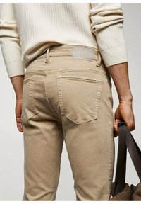 Jeans de mezclilla beige con un diseño ajustado, que cuentan con dos bolsillos traseros y una etiqueta de cuero en la cinturilla. El modelo sostiene una correa marrón.