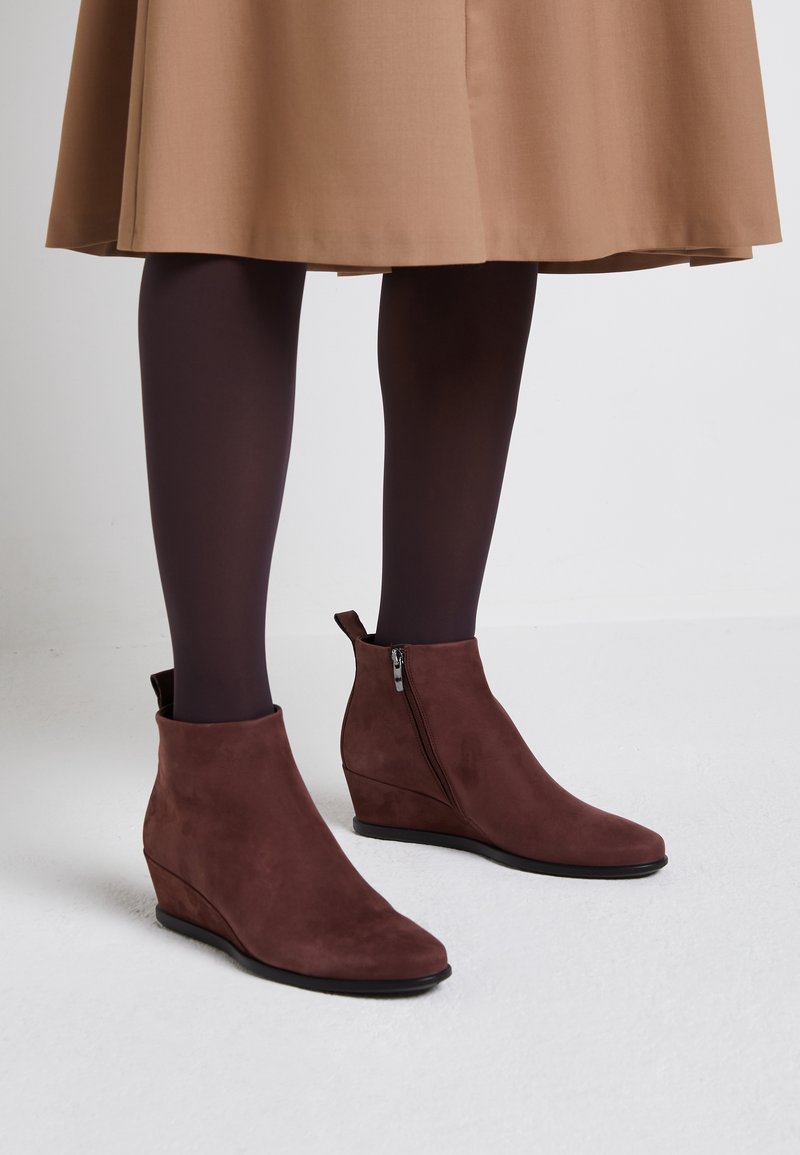 Bottes à talon compensé marron à la cheville avec fermetures éclair latérales, portées avec des collants foncés et une jupe beige jusqu'aux genoux, posées sur un sol blanc.