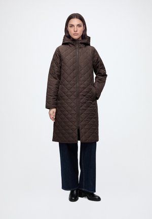 Femme portant un long manteau matelassé marron à capuche avec fermeture éclair devant, pantalon ample foncé et bottines noires, debout devant un fond uni.