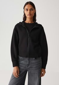 Felpa nera con cappuccio e zip, dal design cropped e con polsini a coste, indossata sopra un top nero a coste e abbinata a jeans grigi.