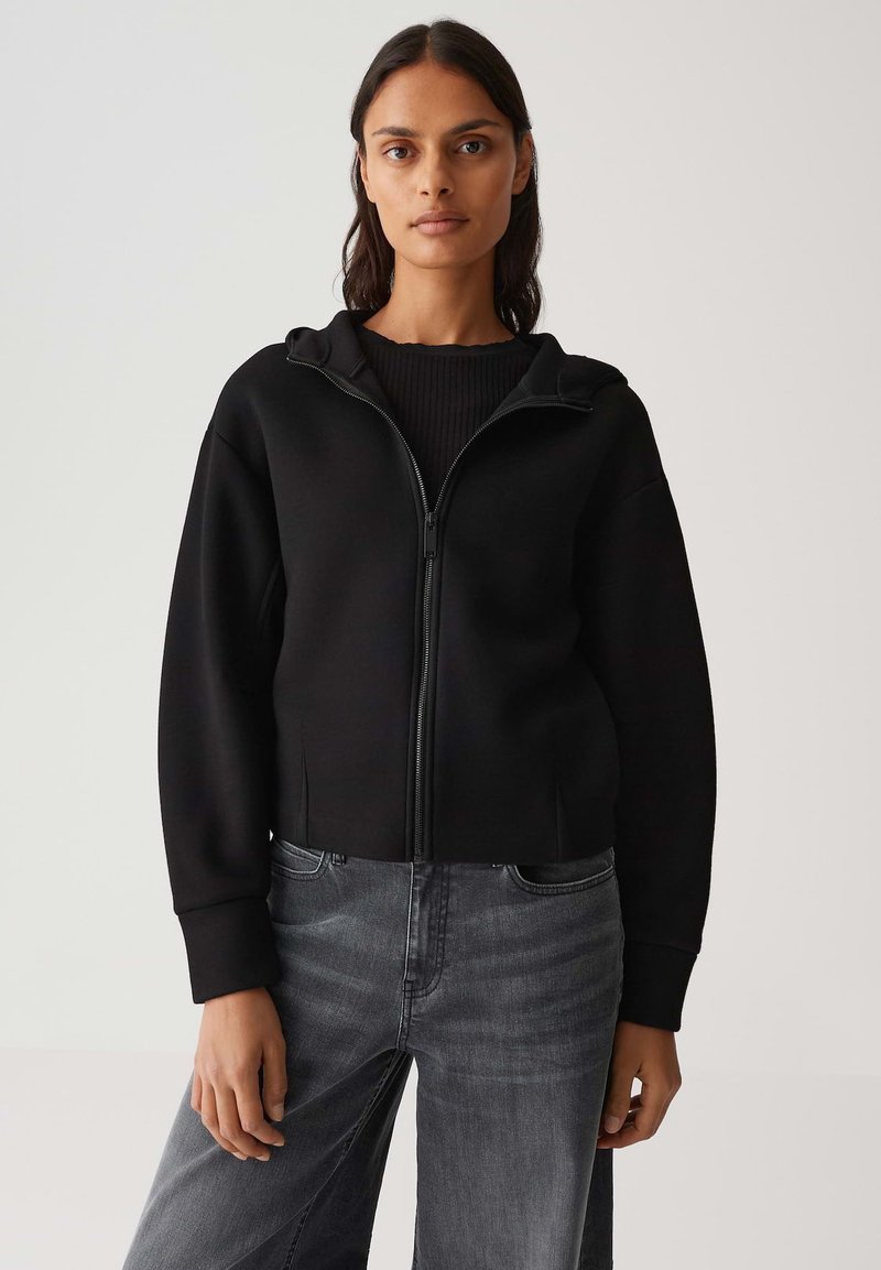 Felpa nera con cappuccio e zip, dal design cropped e con polsini a coste, indossata sopra un top nero a coste e abbinata a jeans grigi.