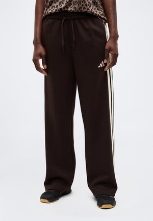 Persona che indossa pantaloni della tuta Adidas neri con strisce laterali bianche e scarpe da ginnastica nere con suola in gomma.