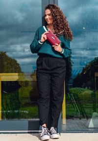 Green knitted sweater with white accents, black pleated trousers, holding a red wallet. Black sneakers with white soles. Outdoor setting.