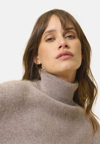 Donna con capelli castano chiaro che indossa un maglione beige a collo alto, che guarda leggermente verso il basso con un'espressione neutra su uno sfondo neutro.