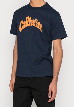 Persona che indossa una maglietta blu navy con il logo arancione acceso "Carhartt" in lettere arcuate, abbinata a pantaloni neri su uno sfondo semplice.