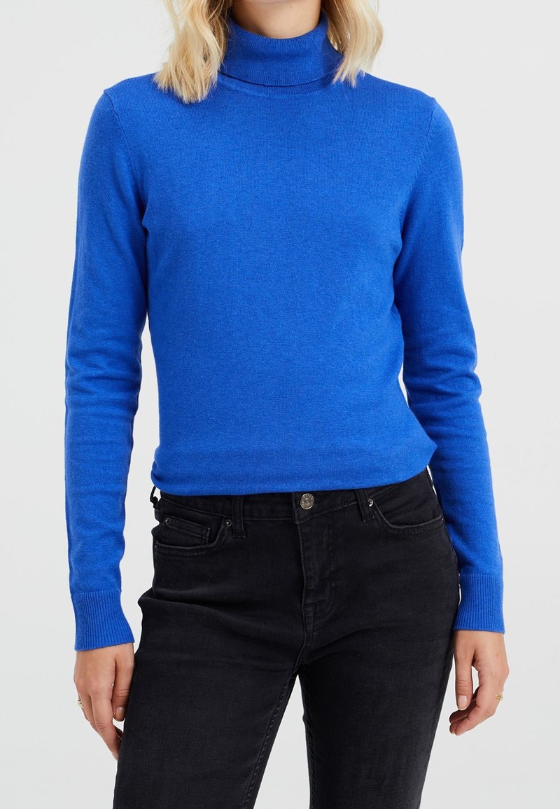Personne portant un pull col roulé ajusté à manches longues bleu vif et un jean noir taille haute devant un fond uni.