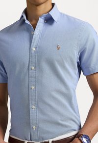 Světle modrá košile s krátkým rukávem, zapínaná na knoflíky, vyrobená z tkané látky, s malým vyšitým logem na levém prsu a bílými knoflíky.