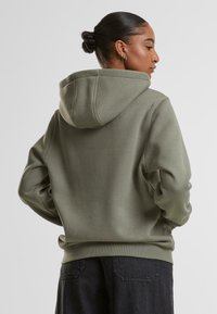 Olivengrønn hettegenser med myk tekstur, med en stor hette, ribbet kant og avslappet passform. Båret med mørke jeans, viser bakvyn.