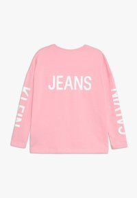 Vaaleanpunainen pitkähihainen paita, jossa takana valkoinen teksti "JEANS" ja vasemmassa hihassa "CALVIN". Pehmeää kangasta ja rento malli.