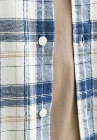 Ein blauer Karobluse mit weißen und beige Akzenten, die eine glatte Textur aufweist und ein kontrastierendes beiges Unterhemd hat.