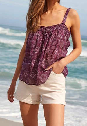 Nainen pukeutuneena violettiin kuvioituun hihattomaan toppiin ja beigenvärisiin käärittyihin shortseihin, seisomassa hiekkarannalla taustalla meren aallot.