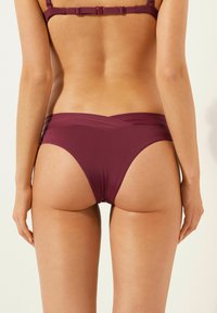 Bas de bikini bordeaux en matériau lisse et extensible. Présente un design à jambe haute avec un dos croisé et une couverture arrière minimale.