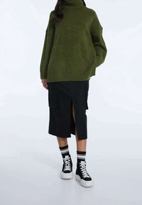 Pull à col roulé vert en tricot, coupe oversize, associé à une jupe cargo noire mi-genou avec une fente latérale, et des baskets montantes noires.