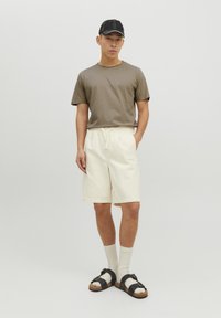 Camiseta en verde oliva, pantalones cortos de algodón crema, gorra negra con costuras blancas, calcetines crema y sandalias negras con detalles de hebilla.