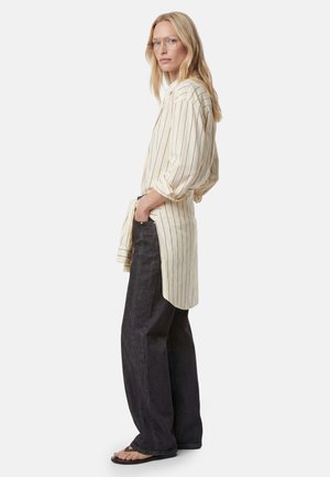 Blond vrouw draagt een oversized beige gestreept overhemd, zwarte wijde jeans, slippers en doorzichtige bril, staat met handen in de zakken.