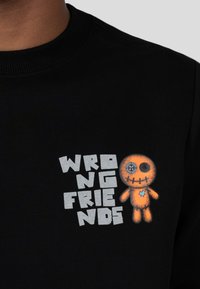 Zwarte shirt met blokletters "WRONG FRIENDS" naast een oranje gestikt poppetje met een patch-oog en een blauw hart op zijn borst.