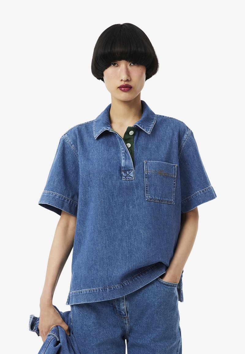 Personne portant une chemise à manches courtes en jean bleu et un jean, tenant une veste en jean assortie, avec une coupe au carré noire et une expression neutre.