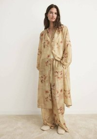 Completo lungo di colore marrone chiaro con motivi floreali; tessuto trasparente; camicia con bottoni dal taglio rilassato; pantaloni abbinati; indossato con calzature beige.