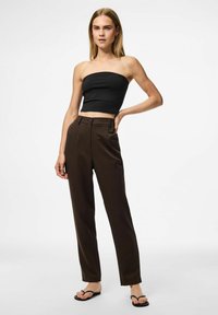 Zwart strapless top gecombineerd met hooggetailleerde bruine broek. De broek heeft een gladde textuur, taps toelopende pijpen en een sluiting met knoop aan de voorkant. Zwarte flip-flops.