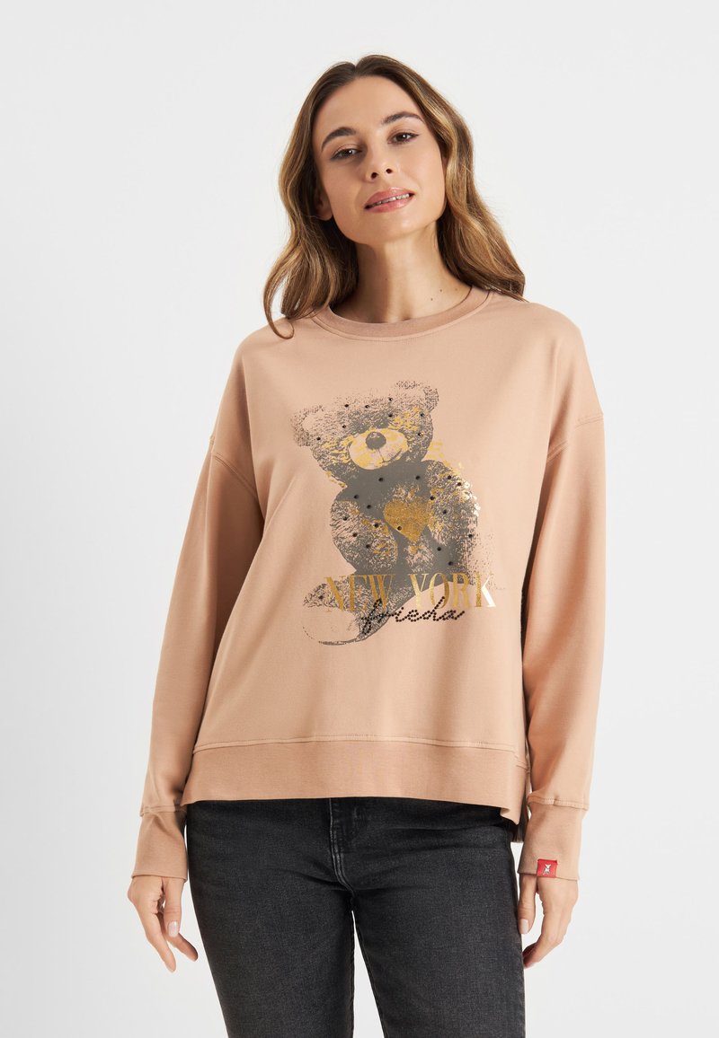 Jasno brzoskwiniowy sweatshirt z grafiką niedźwiedzia i złotym napisem "NEW YORK". Ma okrągły dekolt i ściągacze przy rękawach, noszony z czarnymi dżinsami.