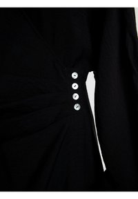 Schwarze Bluse aus strukturiertem Stoff, mit V-Ausschnitt und fünf weißen Knöpfen entlang der Seitennaht. Die Faltenoptik sorgt für visuelles Interesse.