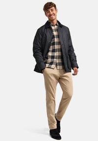 Svart vaxad jacka över en beige- och svartrutig skjorta, i kombination med tanbyxor och svarta stövlar, med en avslappnad passform och knappdetaljer.
