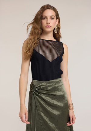 Mujer con cabello largo y ondulado, llevando pendientes dorados, top negro sin mangas con detalle translúcido a rayas y falda verde de satén fruncida.