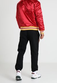 Červená saténová bomber bunda se žlutobílými pruhovanými žebrovanými lemy na rukávech, v kombinaci s černými tepláky a bílými teniskami se zelenými akcenty.