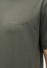Σκούρο ελαιδί πράσινο κοντομάνικο πουκάμισο με ανάγλυφο κείμενο "ARMANI EXCHANGE MILANO / NEW YORK" στο στήθος.